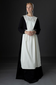 An Amish woman wearing a black dress with a white apron and cap standing against a studio backdrop