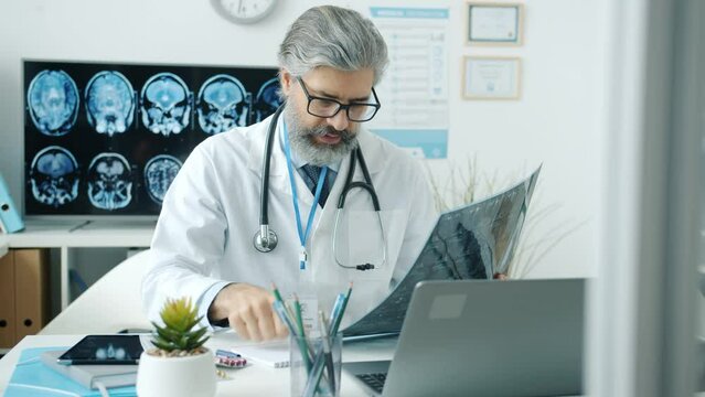 Experienced doctor studying MRI image and using computer typing working in hospital room. Healthcare and modern technology concept.