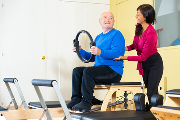 Cheerful Hispanic woman fitness trainer controlling positive elderly man performing exercises with Magic Circle in pilates studio