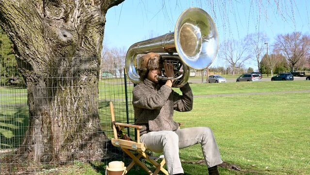 Young man artist with his brown hat plays slow melody on the tuba while sitting on a chair at the park.