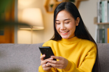 Young Asian beautiful woman sitting on sofa using smartphone, mobile for surfing internet, social media, online shopping, chatting with friends at home