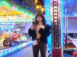 Modern woman in hat using a mobile phone next to a crazy car attraction at night