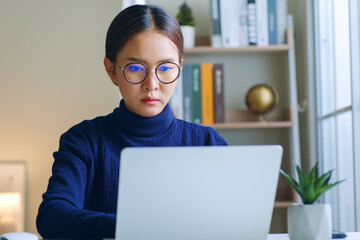Young Asian woman wearing eyeglasses working on laptop seriously at home