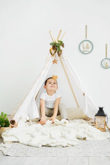 Fototapeta na wymiar Little Caucasian boy is sitting in a toy white wigwam and smiling while looking at a wooden heart. Scandinavian Christmas interior of a children's room.