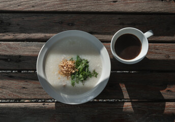 Pork congee or Rice porridge with minced pork sprinkled with deep-fried garlic and coriander in white bowl Served with cup of black coffee on old wooden table for new morning. Asia breakfast, 