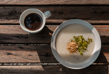 Pork congee or Rice porridge with minced pork sprinkled with deep-fried garlic and coriander in white bowl Served with cup of black coffee on old wooden table for new morning. Asia breakfast, 