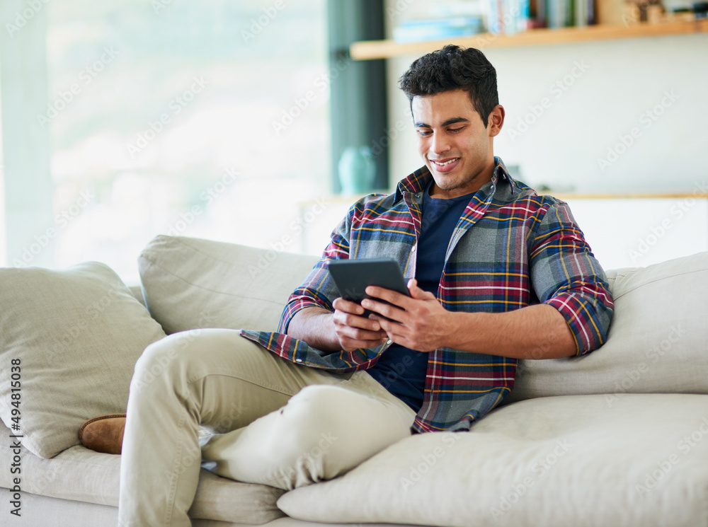 Poster having a little fun online. shot of a handsome young man using his tablet while relaxing on the sofa