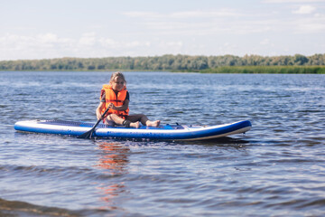 Little blonde girl puddle boarding on lovely lake rowing with oar in hands looking at rippled water in vest life jacket. Active holidays. Inculcation of love for sports from childhood.