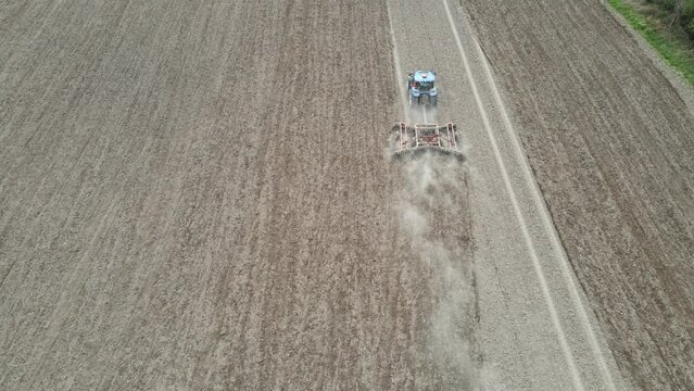 Birds eye view of farmer tilling field in preparation for planting. Evenly spaced rows in the dry soil kicking up dust. Tree line along the edge of the field. Blue tractor in the field. 
