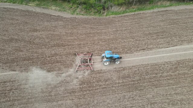 Birds eye view of farmer tilling field in preparation for planting. Evenly spaced rows in the dry soil kicking up dust. Tree line along the edge of the field. Blue tractor in the field. 