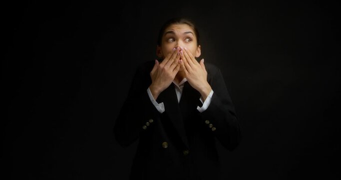 Funny brunette, dressed in formal business clothes, stands on an isolated black background in the studio, covers her mouth with her hands, blurted out something wrong, looks around with her eyes