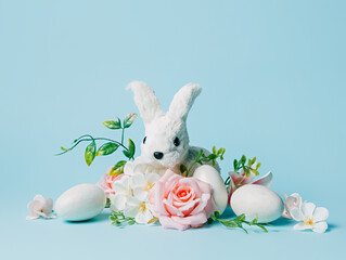 White Easter bunny surrounded with eggs, rose flowers and leaves on pastel blue background. Minimal Easter holiday concept. Creative traditional spring composition. Banner with copy space for text.