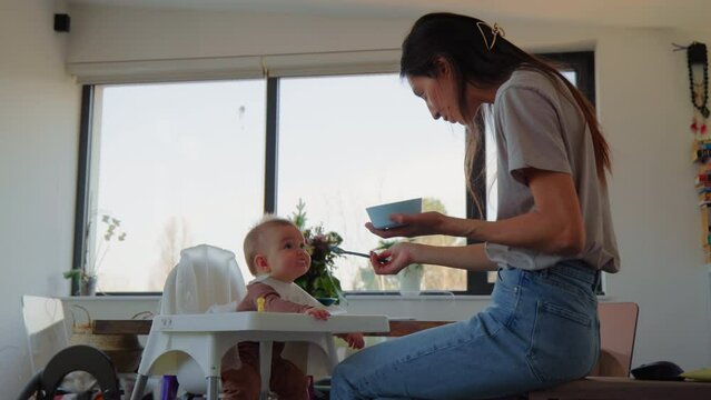 Young mom feeding baby in feeding chair in apartment