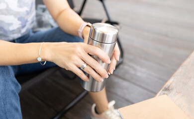 Closeup of woman’s hands hold insulated stainless reusable water bottle sitting in outdoor cafe for takeaway. Bring your own bottle, Eco friendly, zero waste and green living lifestyle.