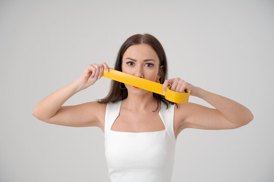 Frightened woman holding tape in front her face covering her mouth and lips. Concept prohibition statements. Caucasian woman is afraid to say her opinion in a white stylish dress on light background