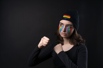 Obraz na płótnie Canvas Portrait young crying woman with painted LGBT flag on her cheek. Crying with rainbow in black hat with rainbow in form heart on black background. Concept violation rights and equality of LGBT people.