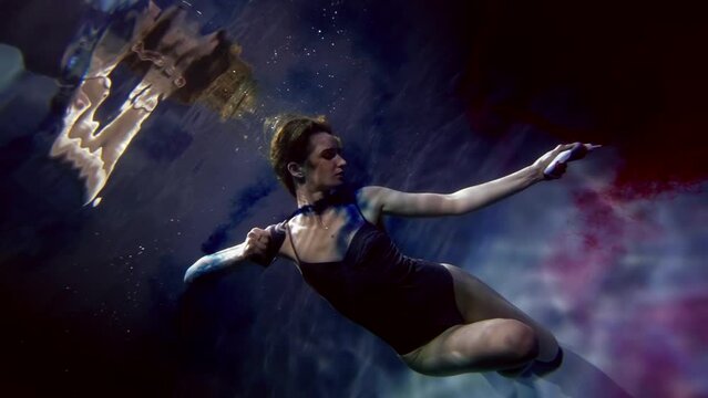 graceful woman is dancing underwater with red and blue colors, painting water of pool, slow motion