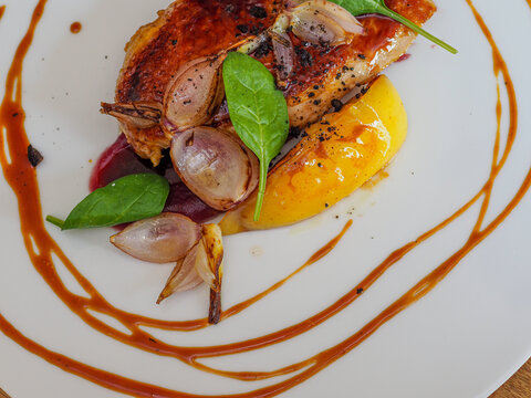 overhead shot of grilled chicken breast with  potatoes, onions, sauces , leafy greens and other vegetables plated in a modern contemporary  way on a white dish over a wooden design table