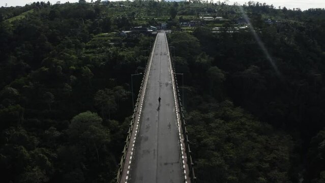 Aerial of male jogging in wilderness running along concrete suspension bridge crossing the green natural forest with mountains peak view at distance, success determination goal concept