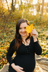 Beautiful pregnant woman in a black dress in the autumn park. Golden autumn. Fallen leaves. Awaiting the birth of a child.