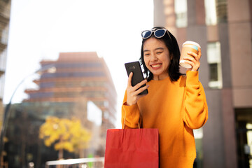 Pretty chinese girl using mobile phone and holding coffee to go. Beautiful woman enjog in coffee while walking on the city street.