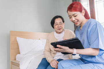 Asian woman nurse wearing scrubs report the health status of Senior Asian woman with digital tablet in the bedroom. Caregiver visit at home. Home health care and nursing home concept.