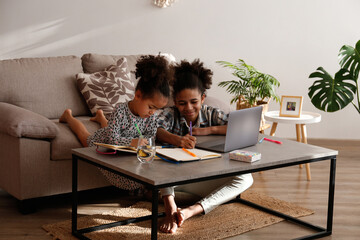 Fototapeta na wymiar Two beautiful black girls of different age on a video call at home. Loving sisters sitting together by the table with laptop. Interior background, close up, copy space.