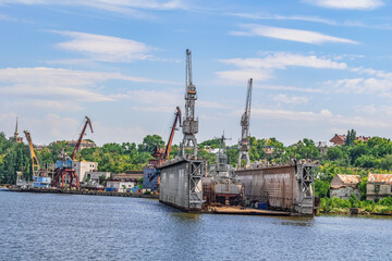 Fototapeta premium Mykolaiv, Ukraine - July 26, 2020: Mykolayiv Shipyard Dock on Inhul River. Coastline with port cranes against the backdrop of green nature