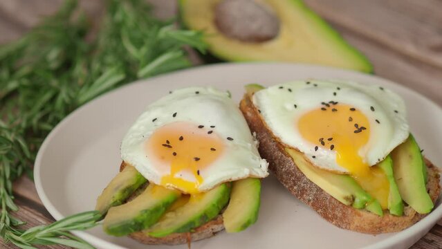 Healthy breakfast for two, couple. Spreading Yolk Two Toasts avocado fried egg, rosemary. 4K Preparing toast breakfast with avocado slices rye wholegrain bread and sunny side up egg on top