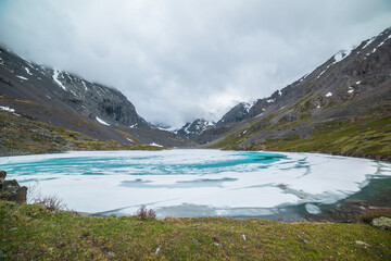 Atmospheric mountain landscape with frozen alpine lake and high snowy mountains. Awesome overcast scenery with icy mountain lake on background of snow mountains in low clouds. Scenic view to ice lake.
