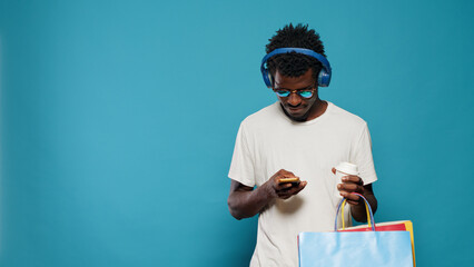 Modern adult using smartphone and carrying shopping bags from retail store. Young man with sunglasses listening to music on headphones and looking at phone after clothing purchase