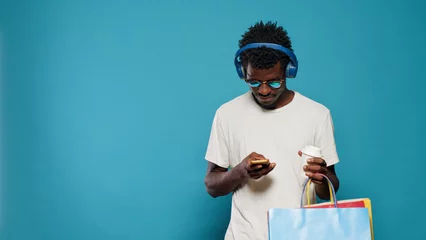 Photo sur Aluminium Magasin de musique Modern adult using smartphone and carrying shopping bags from retail store. Young man with sunglasses listening to music on headphones and looking at phone after clothing purchase
