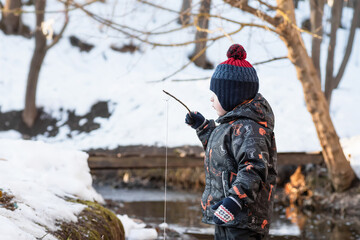 Fototapeta premium A little boy in the park in early spring. A child is playing with a homemade fishing rod made of a stick. Wooden bridge over the stream.