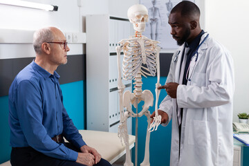 Physiotherapist doctor showing anatomical human skeleton explaining back pain to old man patient...