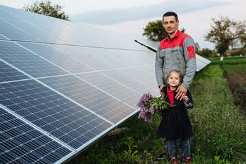 The child hugs the dad the electrician, on a background of solar panels. The engineer shows the...
