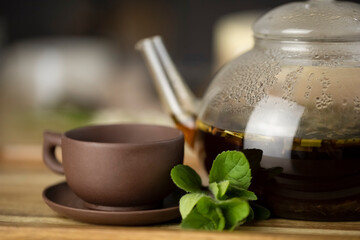 Glass teapot with tea, clay cup and green fresh mint leaves on a wooden table. Tea ceremony and breakfast concept.