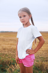 Fototapeta na wymiar Cute little girl in white t-shirts standing in field outdoor. Mock up.