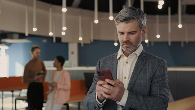 Successful businessman in formal wear usin smartphone standing at the moders office. Text message, reads and views cell phone. Indoor. Application app. Senior people and gadgets concept.