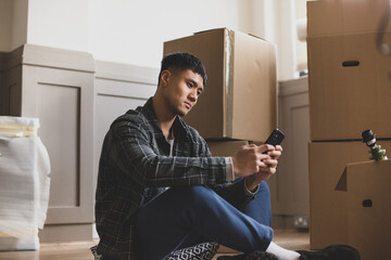 Mid adult male sitting on the floor in new home using smartphone surrounded by boxes