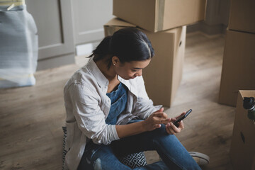 Mid adult female sitting in new home using smartphone surrounded by boxes