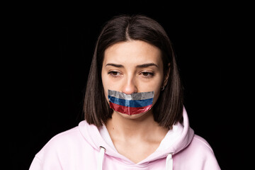 Front camera view. Portrait of young upset girl with three colors duct tape over her mouth isolated on dark background. Censorship, freedom of speech concept.
