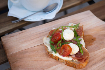Delicious rustic sandwich for comfort breakfast - whole grain bread, prosciutto ham, mozzarella cheese, arugula, tomato and a cup of yummy cappuccino coffee