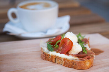 Delicious rustic sandwich for comfort breakfast - whole grain bread, prosciutto ham, mozzarella cheese, arugula, tomato and a cup of yummy cappuccino coffee