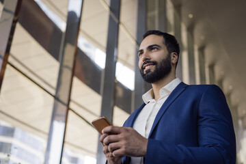 Handsome smiling middle eastern business man using mobile phone working in modern office. Confident arabian manager wearing formal wear text messaging looking at window. Successful business 