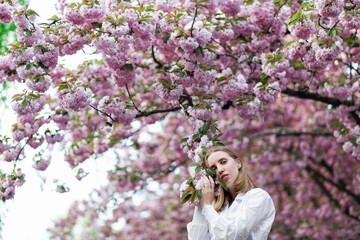 A blonde woman in a white shirt poses near the cherry blossoms. Spring mood