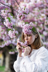 A blonde woman in a white shirt poses near the cherry blossoms. Spring mood