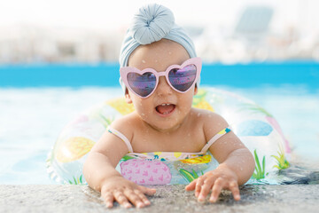 Cute funny toddler girl in colorful swimsuit and sunglasses relaxing on inflatable toy ring...