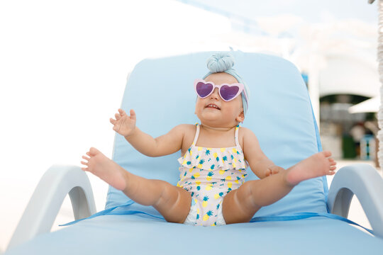 Funny cute baby girl on summer vacation. Child having fun in swimming pool. Sweet toddler girl in colorful swimsuit and sunglasses relaxing on sunbed. Free space for text.