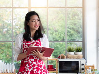 Pretty Asian woman wearing heart shape pattern apron thinking of the menu to cook while holding cookbook in the kitchen.