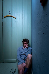 Verwirrter ungepflegter Mann mit Depressionen und Dreitagebart sitzt hilflos und einsam in der Ecke eines Zimmer.  - obrazy, fototapety, plakaty
